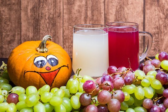 Glass of red stum and 2 glasses of white stum together with some green and red grapes and a funny pumpkin photographed in front of a wooden wall.
