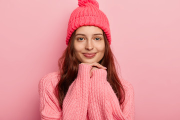 Beautiful young dark haired woman keeps hands uder chin, wears warm winter clothes, looks gladfully at camera has natural beauty with no makeup. Pretty girl in knitted hat and sweater isolated on pink