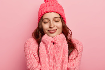 Headshot of satisfied European woman has healthy skin, keeps eyes shut, hands under chin, wears warm hat and oversized sweater, isolated over pink background. People, fashion and good feelings