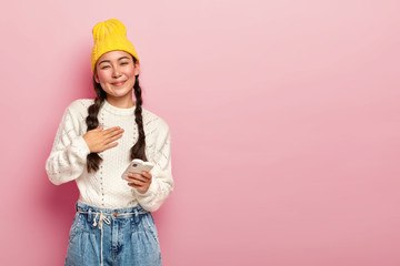 Portrait of pleased Korean woman keeps palm on chest, being touched by heartwarming words, holds modern electronic device, receives good message, wears yellow hat, casual white jumper and jeans