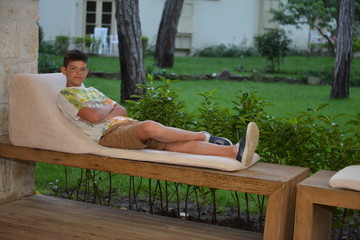 Boy lying on the bench outdoors in the evening. Portrait of teen boy with dark hair. Charming young male in fashionable clothes relaxing on bench in park. Weekend concept.