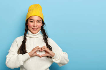 Pretty brunette girl makes heart hand sign, smiles pleasantly, wears yellow hat and white sweater, expresses love and affection, has satisfied expression, models against blue background, copy space