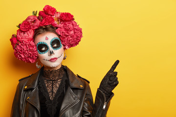 Face art and traditional Mexican holiday concept. Serious young lady wears dead mask skull makeup, flower wreath with strong scent to guide souls from cemetries, points on free space over yellow wall