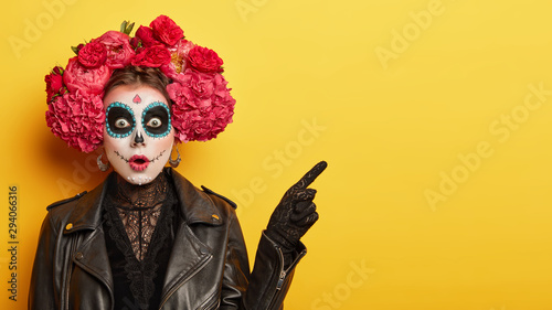 Happy Halloween concept. Scared horrible female in special outfit, wears white clay skull, professional makeup to look spooky, wears black clothes points away on blank space against yellow background