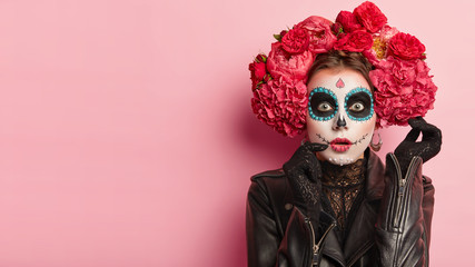 Studio shot of lovely woman wears halloween makeup, dressed in black outfit, red wreath, has zombie...