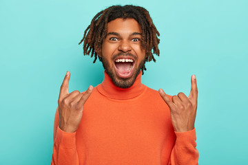 Energized dark skinned young man rocks on party, brings positive vibes, shows rock n roll gesture,...