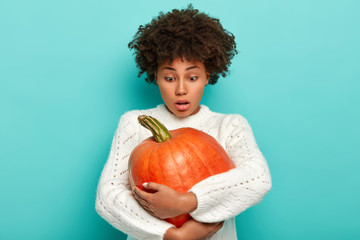 Photo of frightened dark skinned female carries pumpkin for Halloween, focused down with...