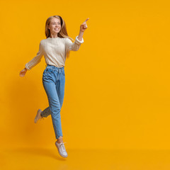 Cheerful teen girl jumping and pointing aside on orange background
