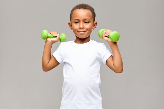 Portrait of cheerful Afro American boy with skinny arms smiling happily at camera while exercising in gym with two dumbbells, going to build strong healthy athletic body. Fitness and children