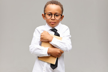 Portrait of handsome dark skinned schoolboy with short Afro haircut posing isolated in studio in glasses, shirt and tie embracing copybook, feeling shy in new school. Learning and knowledge