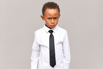 Horizontal image of frustrated dark skinned pupil in school uniform having angry facial expression, frowning, being mad at rude classmate bully, ready to stand for himself, posing in studio