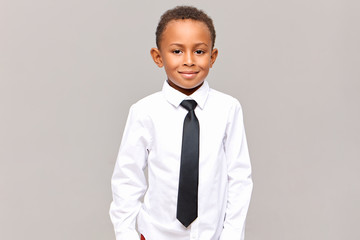 Waist up studio shot of handsome neat dark skinned male elementary pupil posing isolated dressed in white clean ironed shirt and black elegant tie, ready for going to school, smiling at camera
