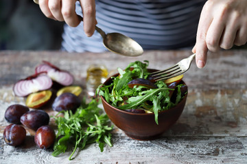 Male hands mix salad in a bowl with spoon and fork. Light salad with arugula and plums. Autumn plum salad. Healthy food. Selective focus. Macro.