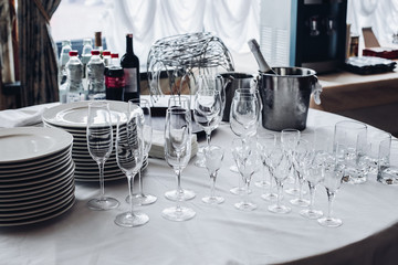 View over white round table with stacks of white plates, empty champagne flutes and glasses with bottles of water and ice buckets.