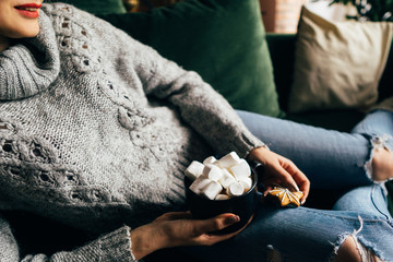 Christmas, cold autumn or winter day. Warming mood. Woman drinking warm cocoa with marshmallows. Lazy weekend in knitted sweater on the couch. Cozy scene, hygge concept