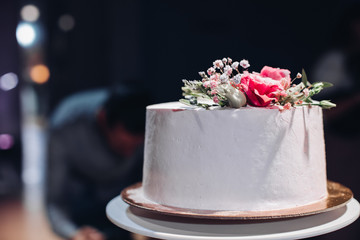Close-up of beautiful white smooth wedding cake decorated with dried flowers on the top in artificial light.