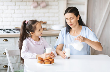 Mother pouring milk to glass, sitting in kitchen with daughter