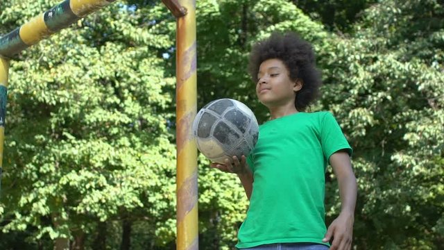 Smiling black school pupil playing ball standing stadium gate, sport activity