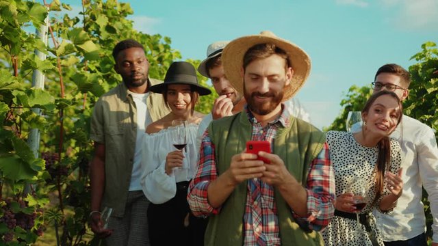 Smiling farmer in strawhat using smarthone application at vineyard. His curious friends looking on mobile phone laughing in behind.