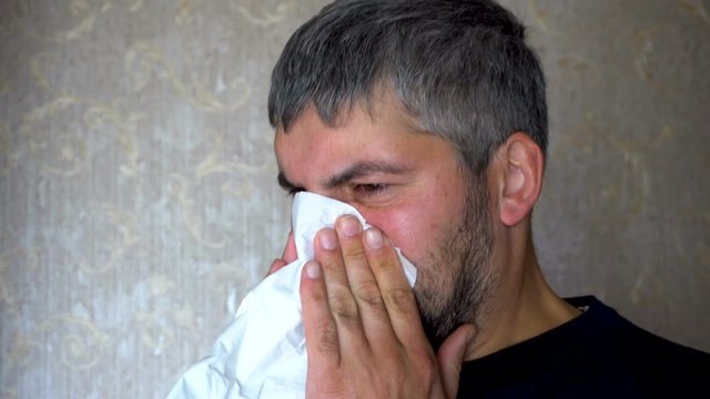 Sick man blows his nose in a paper handkerchief. Unshaven face. Allergic reaction. Unhealthy look. Heat. Dark clothes. Against the background of the interior of the house. Close-up.