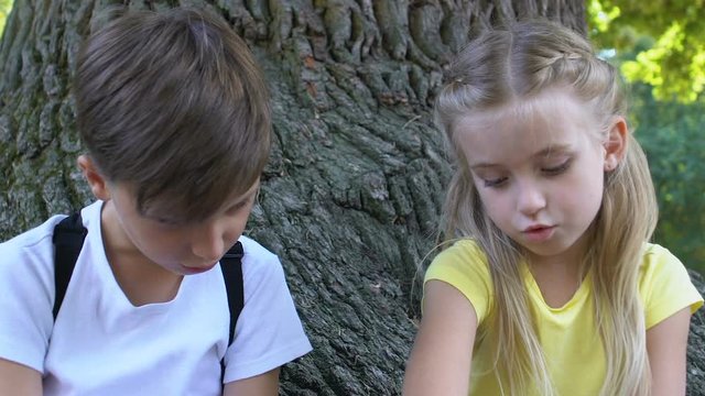 Cute male and female kid talking sitting in garden under tree, communication