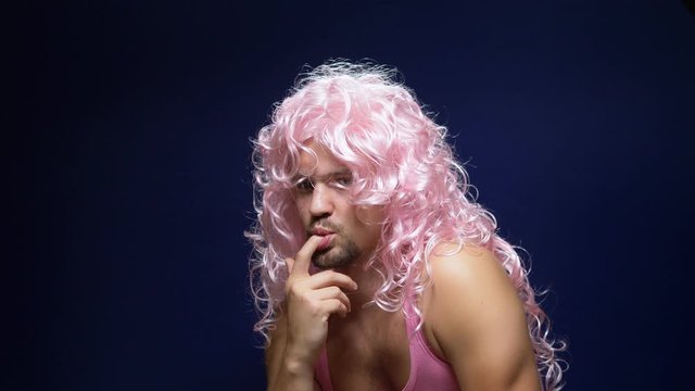 crazy handsome young guy in a curly wig and a pink t-shirt against a dark background is dancing funny, shows his muscles