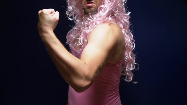 crazy handsome young guy in a curly wig and a pink t-shirt against a dark background is dancing funny, shows his muscles