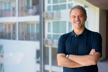 Happy mature handsome Hispanic man smiling with arms crossed by the glass window at home