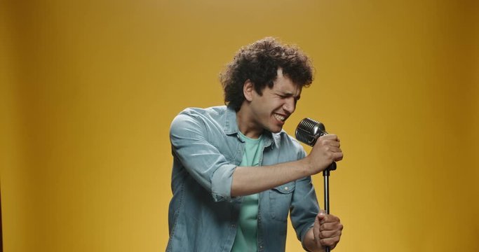 Close up shot of asian man with curly hair in casual clothes crazily dancing and singing in microphone, isolated on yellow background 4k