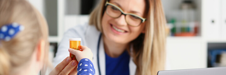 Beautiful smiling female doctor hold in arms pill bottle and offer it to child visitor closeup....