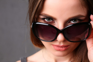 Naklejka premium Beautiful young smiling brunette girl wearing sunglasses winking portrait over grey background. Modern sly coquette lady killer hunter plan concept