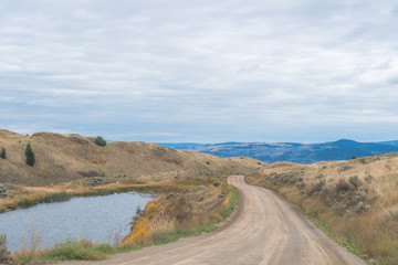 Dirt road in the valley of desert hills with shrub, dry trees, pine trees, mountain range in the background, rural countryside area, small pond near Kamloops