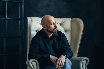 sad miserable serious middle-aged man in a black shirt sits and thinks about his problems in a room...
