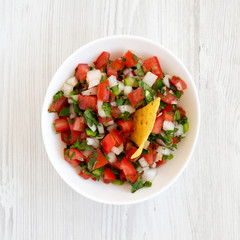 Pico de Gallo in a white bowl on a white wooden table, top view. Overhead, from above, flat lay. Close-up.