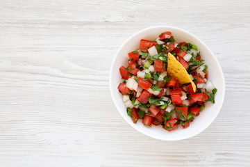 Pico de Gallo in a white bowl on a white wooden surface, top view. Overhead, from above, flat lay....