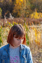 A girl stands with her head bowed on a sunny day against the backdrop of an autumn forest. The girl has red hair. Wood and reeds in the background is blurred.