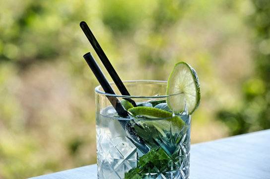 Glass with a non-alcoholic refreshment with lemon and mint on a table on a terrace against a background with soft light green bokeh