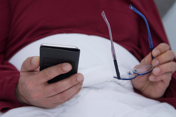 man lying in bed with the cell phone in one hand and the glasses in the other