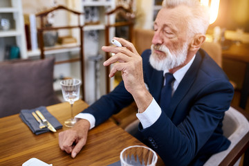 Elegant senior man in suite smoke heat-not-burn tobacco product technology sit in restaurant, holding cigarette in right hand