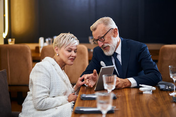 Beautiful senior couple looking at the same tablet in restaurant, public concept, caucasians mature...