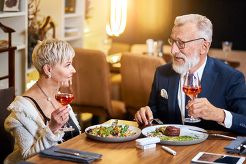 Stylish, elegant senior couple look at each other and chatting, close relationship. Grey-haired man in tuxedo raise a glass. Use e-cigarette, vape. Modern technologies