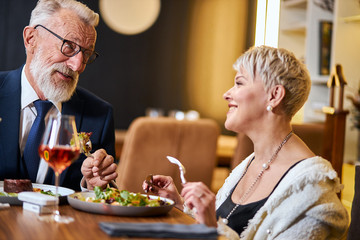 Elderly elegant people taking meal in beautiful restaurant. Free and modern, using iqos, e-cigarettes. Caucasian grey-haired and beared male in tuxedo and glasses. Woman looks at him and listen