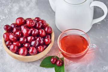 White teapot, herbal tea in glass cup and fresh cherry in wooden bowl on a gray background
