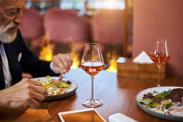 Bearded gray-haired man in elegant restaurant.Close photo of mature hands. Glass of champagne and dish of salad on table.