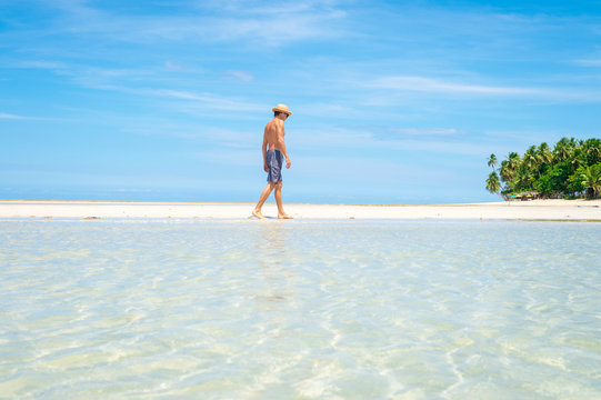 A tourist wearing a straw sun hat walking along a sand bar above the calm shallow waters of a sunny palm-lined tropical island beach 