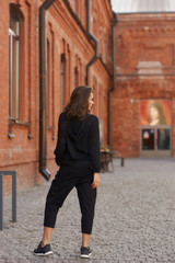 Fototapeta na wymiar Outdoor summer portrait. Young elegant woman in black hoodie and trousers standing and posing at urban city street