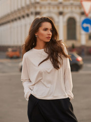 Outdoor summer portrait. Young stylish woman in white hoodie and black trousers standing and posing at city street at sunset