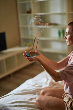 close-up hands of caucasian girl in pajamas meditating at morning while sitting on bed with burning aroma incense sticks in hands. Aromatherapy. Meditation, yoga concept