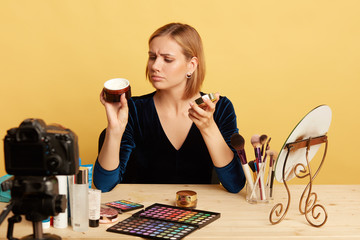 Portrait of glamourous blonde female cosmetics expert looking at new face cream with thoughtful expression, reading attentively annotation on the tube, recording video for beauty blog.