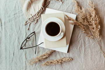 Autumn, fall composition. A cup of coffee lying on the grey linen bed with beige warm blanket, books, glasses and reeds. Lifestyle, still life concept. Flat lay, top view.